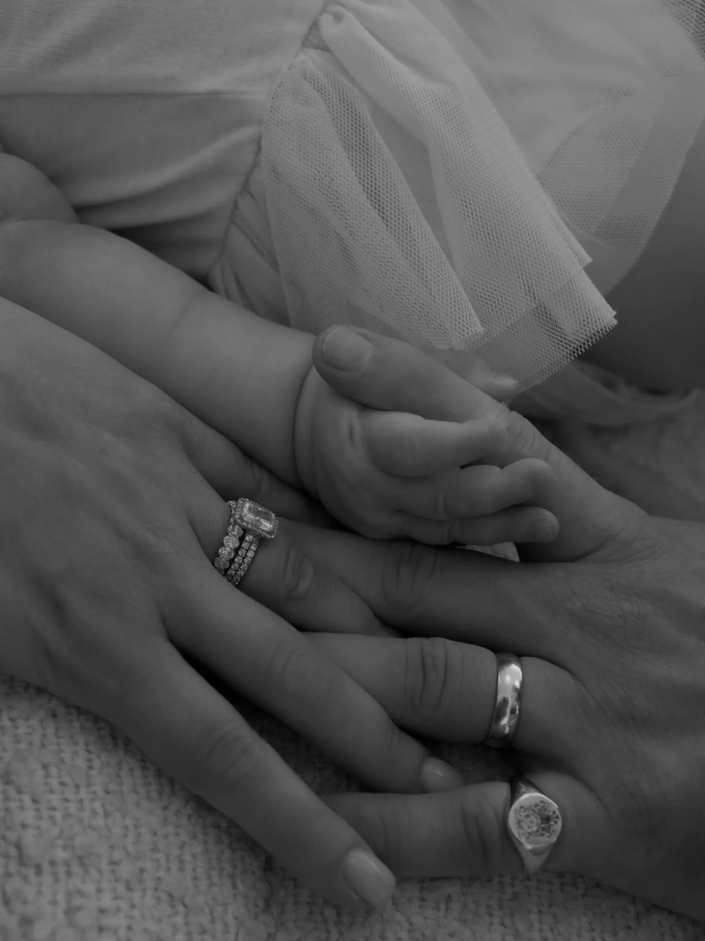 Black and white close-up of a newborn baby's hands resting with their parents' hands and rings