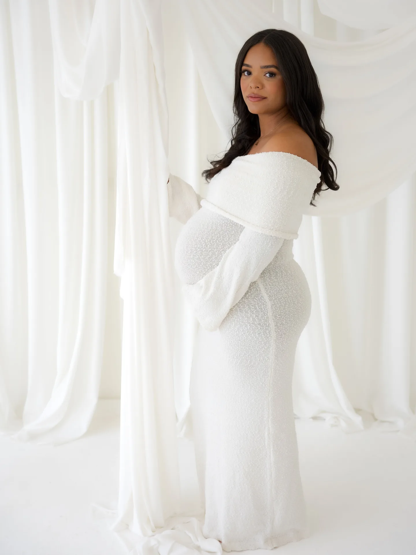 Expectant mother standing in a white gown in a softly lit cream studio