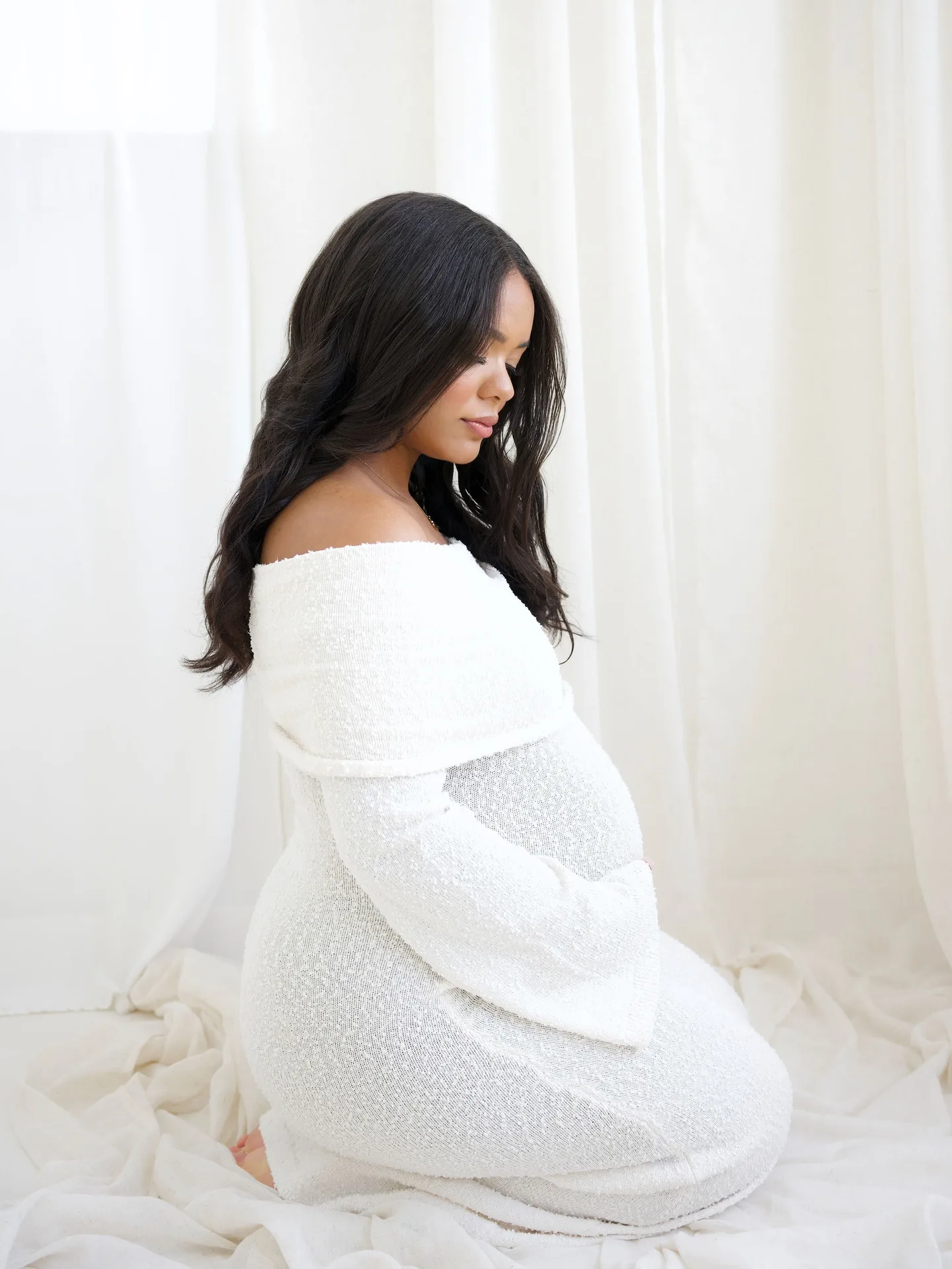 Expectant mother kneeling in a white gown during a soft studio maternity session