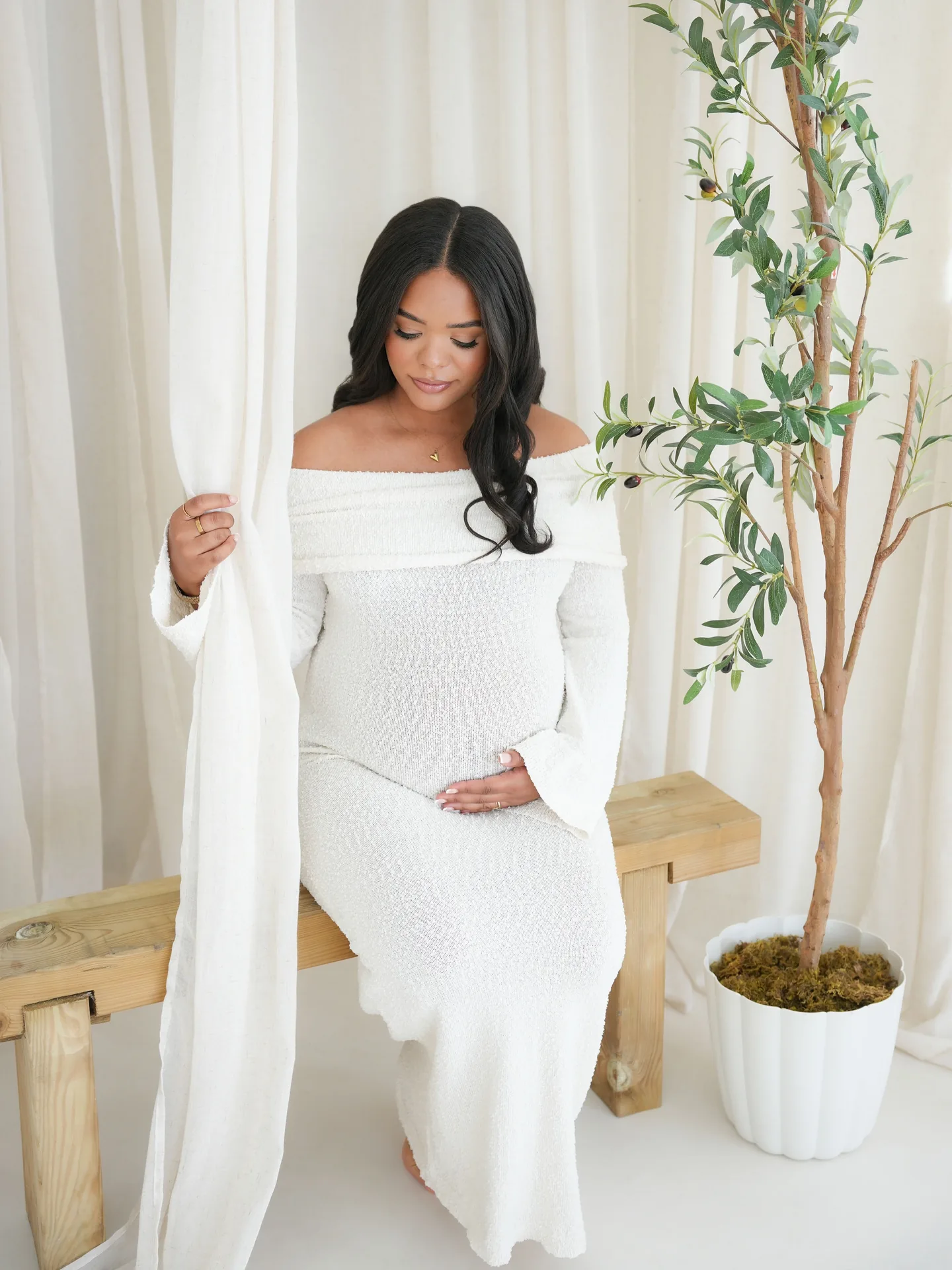Expectant mother seated in a white gown beside a leafy tree in the studio